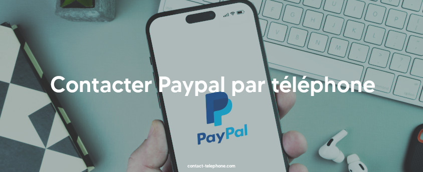 Smartphone affichant le logo de Paypal, il est tenu devant un clavier d'ordinateur et des écouteurs posés sur un bureau.